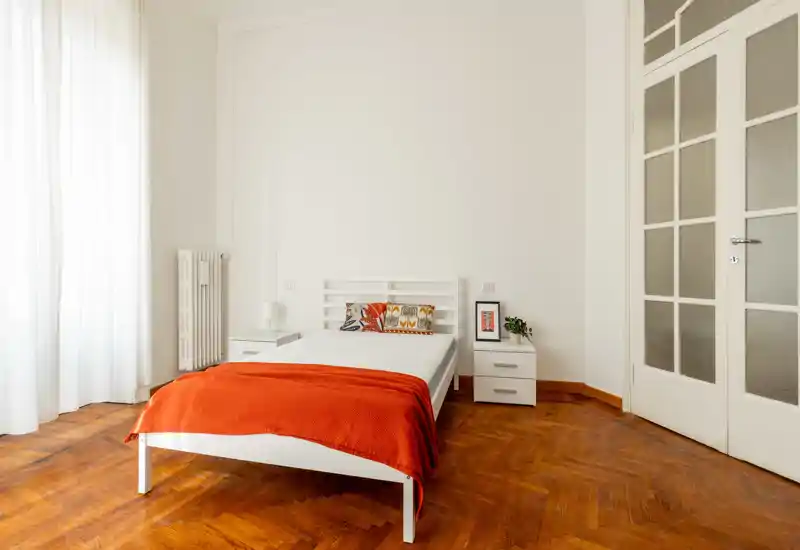 Dormitorio luminoso y minimalista con cama blanca, suelo de parquet cálido y una manta naranja intensa. Gran ventana que aporta luz natural; mesitas y decoración sencilla completan el espacio.