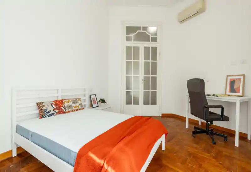 Dormitorio luminoso y minimalista con estructura de cama blanca, suelo de parquet cálido y una manta naranja que aporta color; escritorio y silla para una zona de trabajo pequeña.