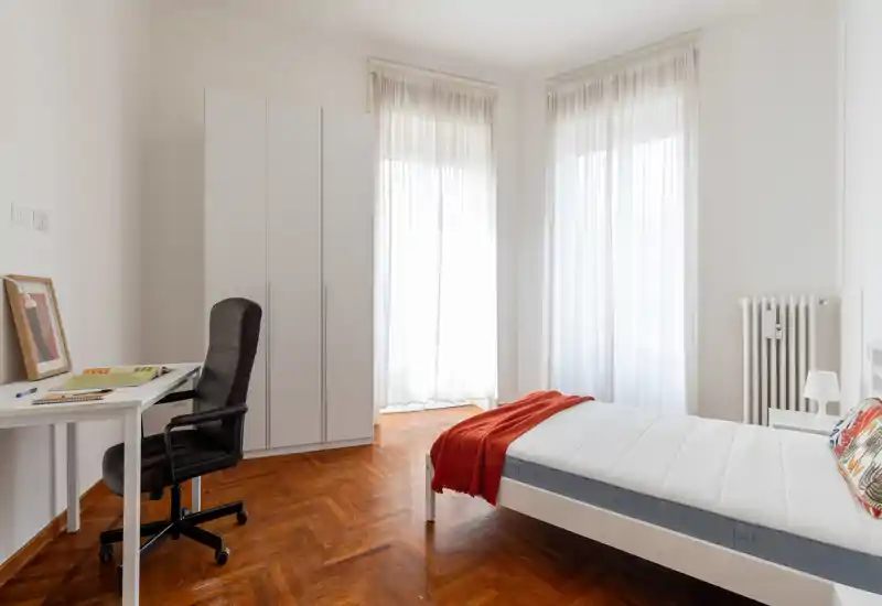 Dormitorio luminoso y minimalista con cama individual con ropa blanca y una manta roja, suelo de parquet y un pequeño escritorio junto a la ventana: ideal para un alquiler cómodo y bien iluminado.