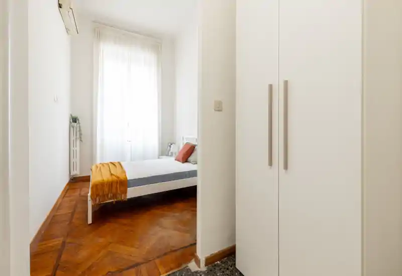 Camera da letto luminosa e minimal con letto bianco, parquet in legno caldo e ampia finestra con tenda velata che diffonde luce naturale.