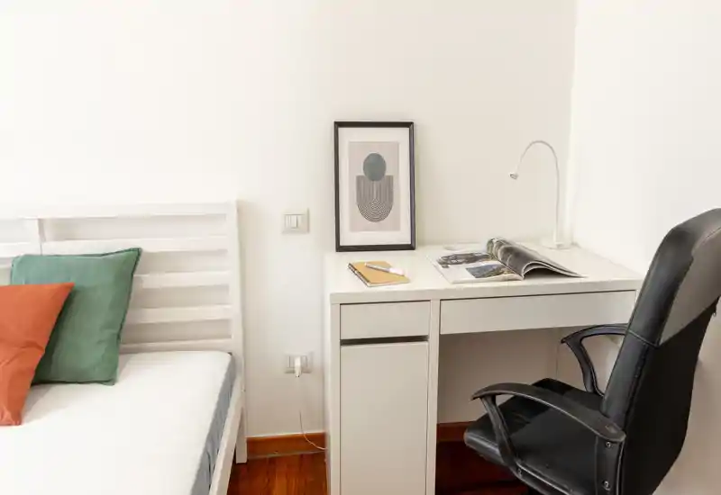 Angolo camera luminoso e minimal con struttura del letto bianca, cuscini colorati e una scrivania bianca ordinata con lampada e sedia da ufficio — ideale per una persona o uno studente.