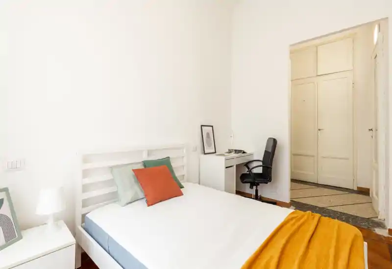 Camera da letto luminosa e minimale con letto in legno bianco, cuscini colorati e una calda coperta arancione; piccolo scrittoio con sedia vicino alla porta per un angolo lavoro pratico.