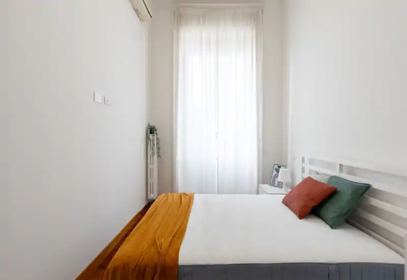 Camera da letto luminosa e minimale con letto matrimoniale ben rifatto, plaid e cuscini colorati, comodino con lampada e tende leggere che diffondono luce naturale morbida.