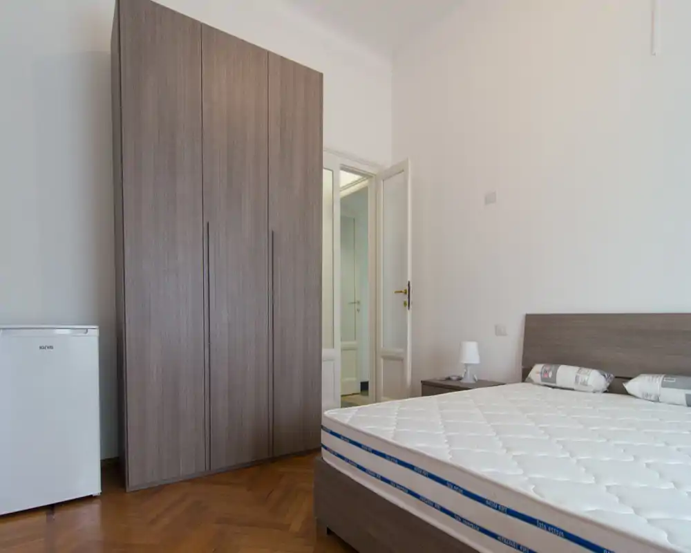 Camera da letto luminosa e minimal con un grande materasso, testiera in legno e armadio coordinato. Pavimento in parquet pulito e comodino con lampada completano lo stile moderno e semplice — perfetta per annunci di affitto incentrati su spazio e luce.