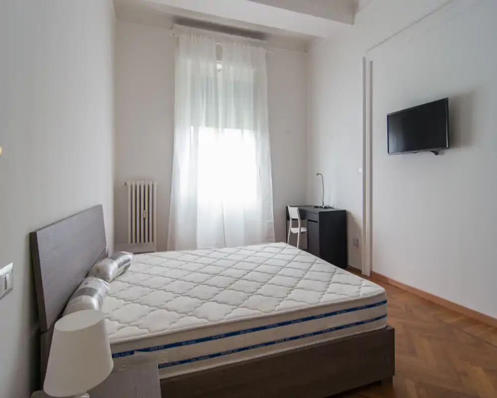 Camera da letto luminosa e minimalista con ampio letto, pavimento in parquet e TV a parete. Una piccola scrivania vicino alla finestra offre un angolo lavoro e tende leggere diffondono la luce.