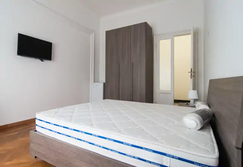 Camera da letto moderna e luminosa con un ampio materasso su una struttura in legno, un armadio alto e una TV a parete; stile pulito e luce naturale abbondante.