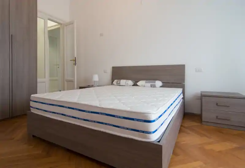 Camera da letto moderna e spaziosa con grande materasso su struttura in legno, comodino e armadio coordinati e pavimento in parquet caldo. Arredamento chiaro e neutro che trasmette un'atmosfera pulita e riposante, perfetta per annunci.