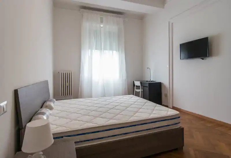 Camera da letto luminosa e minimal con letto ampio, testiera in legno, lampada da comodino e una piccola scrivania vicino alla finestra. Arredi neutri e luce naturale creano un ambiente tranquillo ideale per gli annunci.