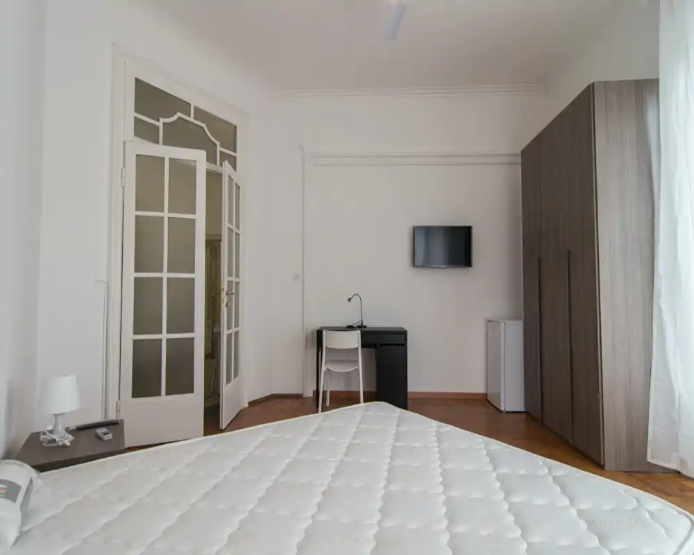 Dormitorio luminoso y minimalista con un gran colchón en primer plano, un armario y un escritorio en la pared del fondo, y luz natural que entra por la ventana a la derecha.