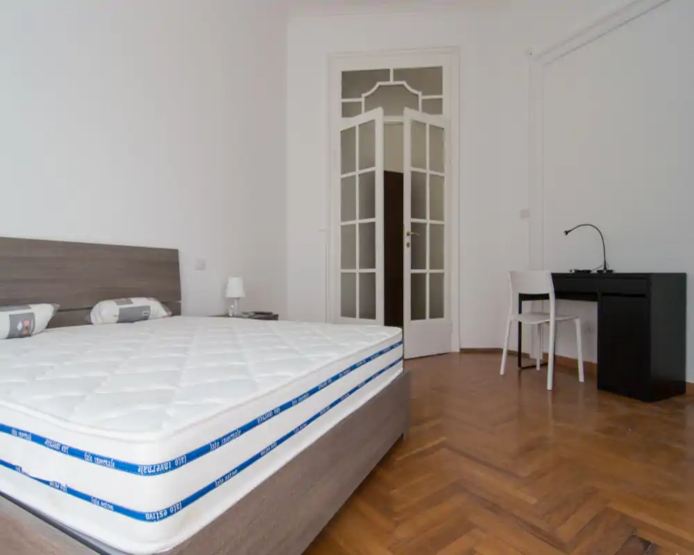 Dormitorio luminoso y minimalista con una gran cama en primer plano, suelos de parquet y un pequeño escritorio en la esquina — ideal para inquilinos individuales o parejas.