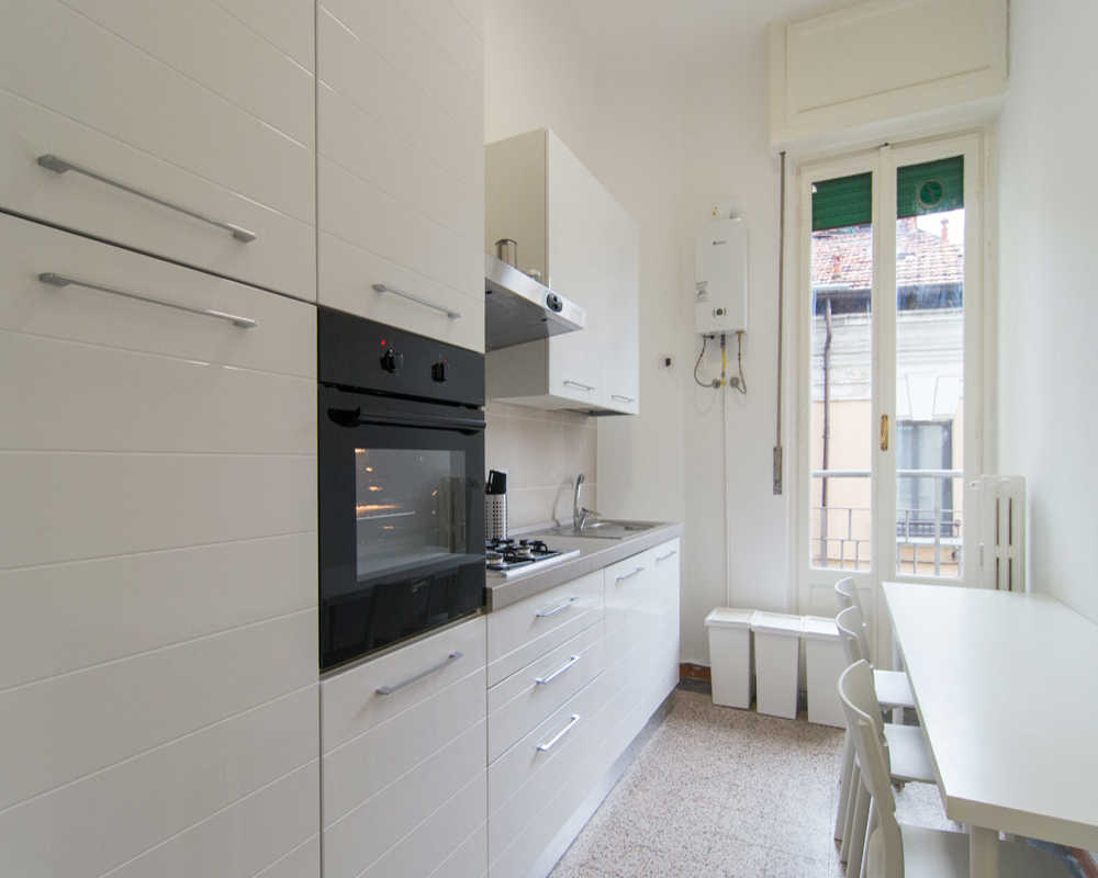 Cucina a corridoio luminosa e moderna con forno integrato, piano cottura a gas e un piccolo tavolo vicino alla finestra. Pensili bianchi e luce naturale creano uno spazio di cottura funzionale e pulito.