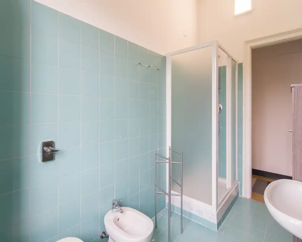 Bagno piastrellato luminoso con cabina doccia, bidet e lavabo; design semplice e pulito con luce naturale tenue.