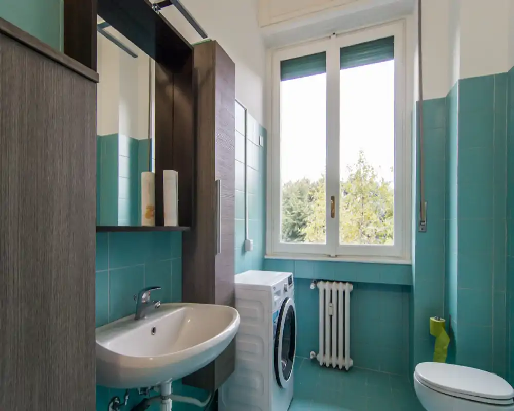 Bagno luminoso e curato con piastrelle turchesi, lavabo con specchio e mobile, lavatrice sotto la finestra e WC; luce naturale da una grande finestra con vista sugli alberi.