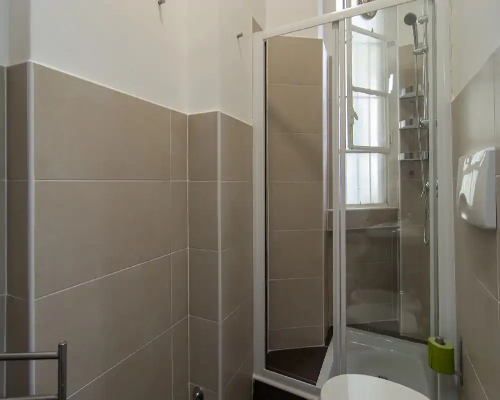 Bagno piastrellato compatto con box doccia in vetro, sanitari a parete e una piccola finestra satinata che fornisce luce naturale.