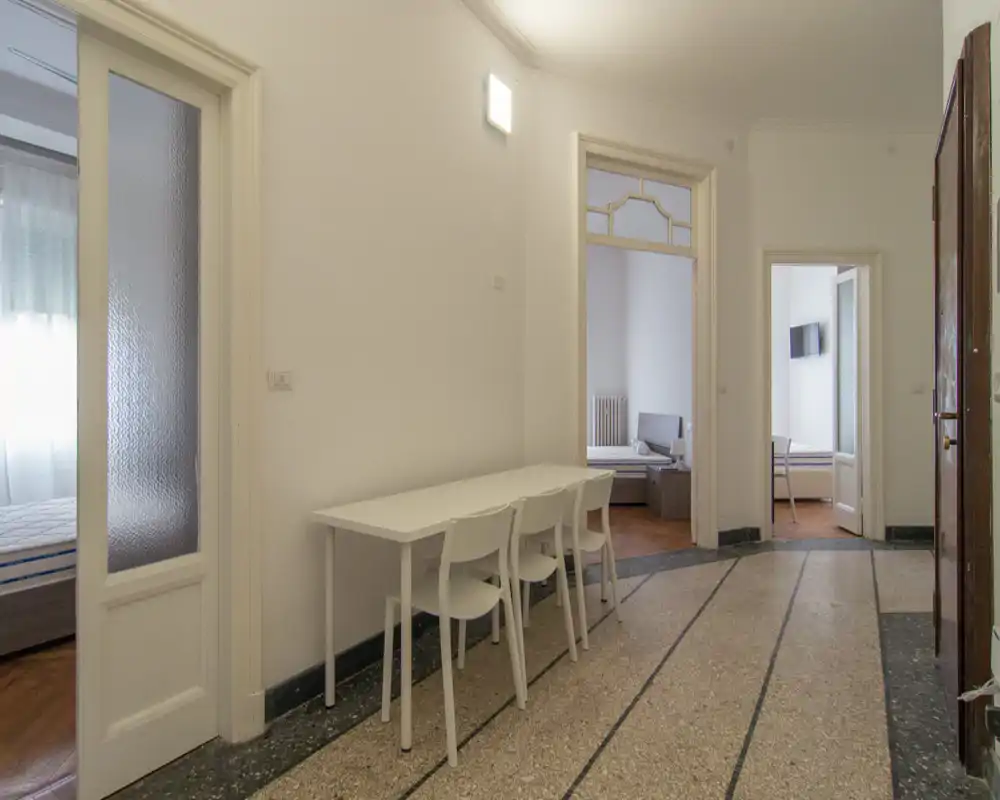 Corridoio d'ingresso con un tavolo e sedie semplici, pareti bianche e porte che conducono alle stanze; luce naturale intensa proveniente dalla sinistra.