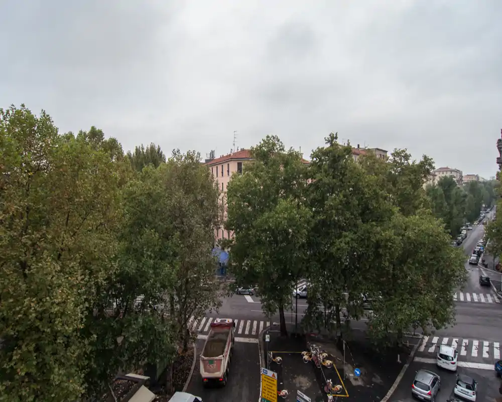 Veduta esterna verso la strada da un piano alto con viale alberato, strisce pedonali e auto parcheggiate sotto un cielo nuvoloso.
