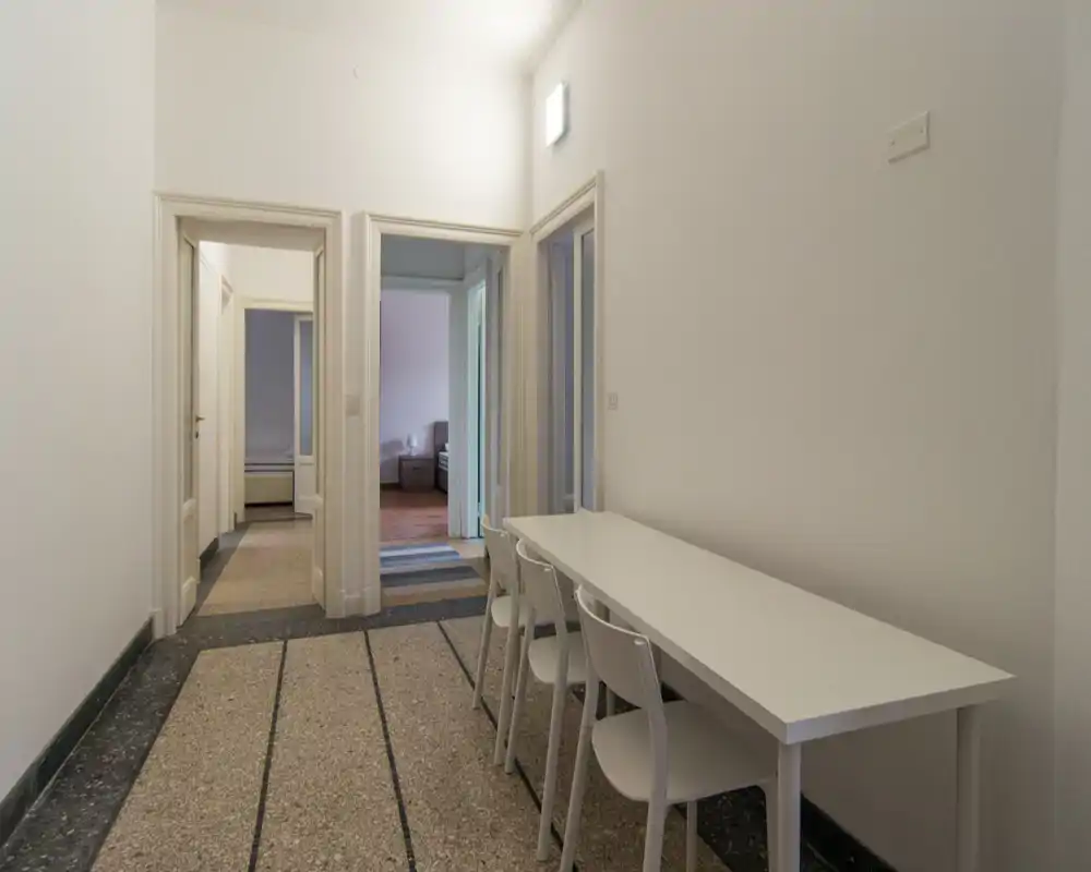 Zona pranzo stretta con un lungo tavolo bianco e quattro sedie, che si apre su stanze adiacenti. Stile semplice e colori neutri, ideale per annunci funzionali.