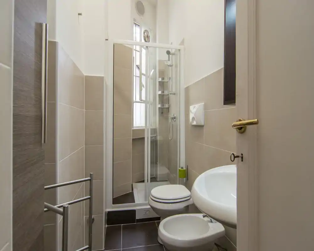 Bagno compatto e luminoso con cabina doccia angolare, wc sospeso, bidet e lavabo; piastrelle beige neutre e una finestra che porta luce naturale.