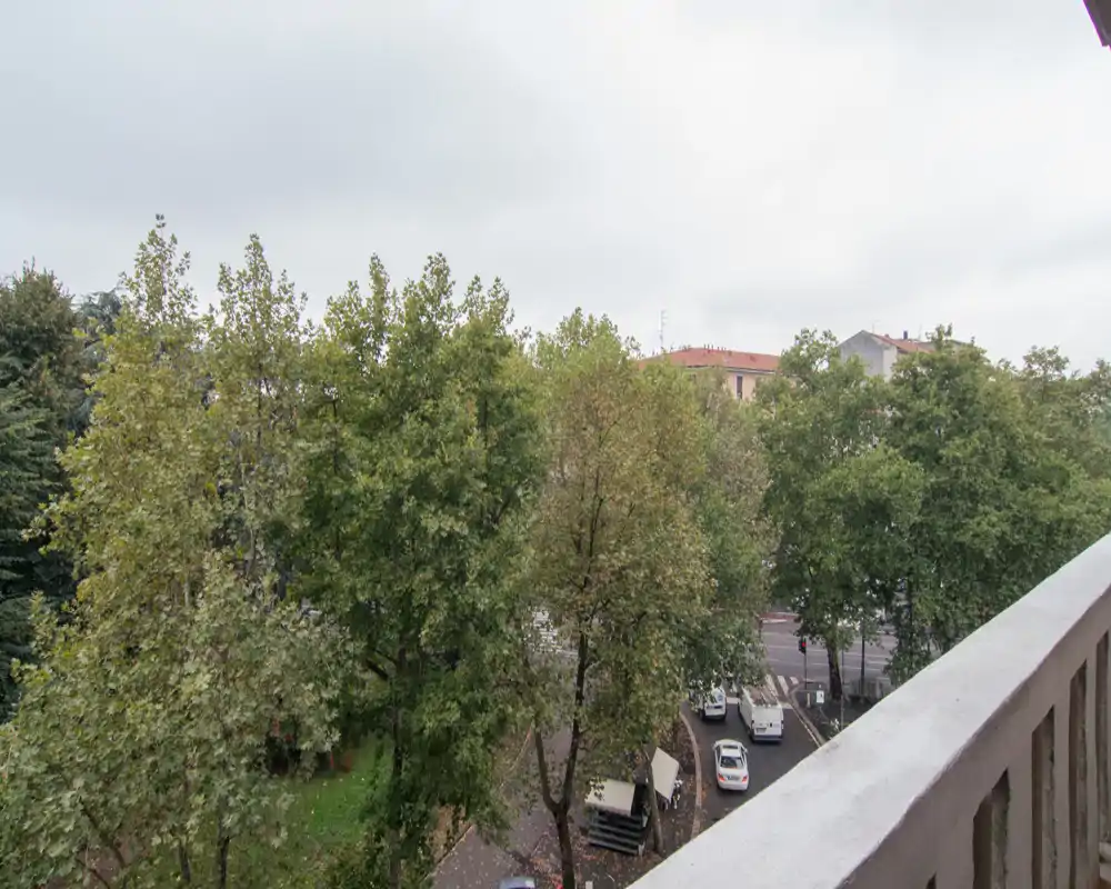 Vista dal balcone con alberi frondosi e una strada sottostante, ringhiera in cemento in primo piano e palazzi sullo sfondo; luce naturale nuvolosa.