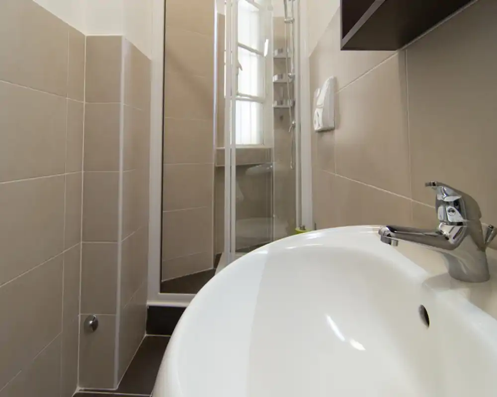Bagno moderno e pulito con primo piano di un lavabo bianco e rubinetto cromato, pareti piastrellate beige e box doccia visibile sullo sfondo.
