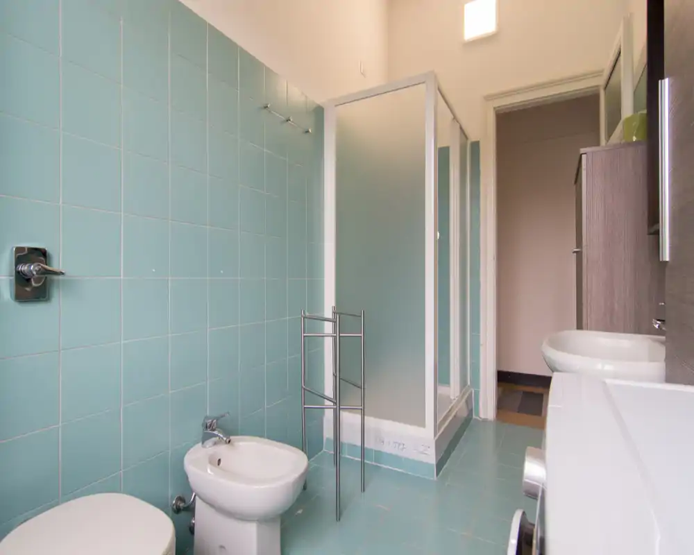 Bagno luminoso e pulito con piastrelle azzurre, bidet e WC, box doccia con vetro satinato e lavabo moderno con mobile.