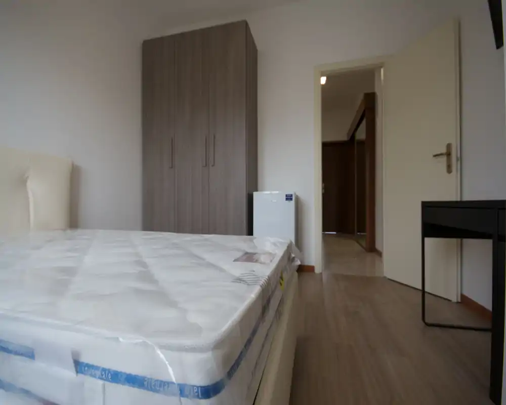 Dormitorio luminoso y minimalista con un gran colchón en primer plano, un armario contra la pared y un pequeño electrodoméstico junto a la puerta. Líneas limpias y tonos neutros ofrecen una sensación moderna y tranquila, adecuada para anuncios de alquiler.