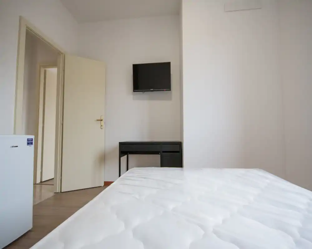 Dormitorio sencillo y minimalista con el colchón en primer plano, un pequeño escritorio y TV en la pared. Paredes blancas y suelo neutro que proporcionan una sensación luminosa y despejada, ideal para alquileres cortos.