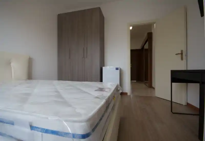 Dormitorio luminoso y minimalista con un gran colchón en primer plano, un armario contra la pared y un pequeño electrodoméstico junto a la puerta. Líneas limpias y tonos neutros ofrecen una sensación moderna y tranquila, adecuada para anuncios de alquiler.