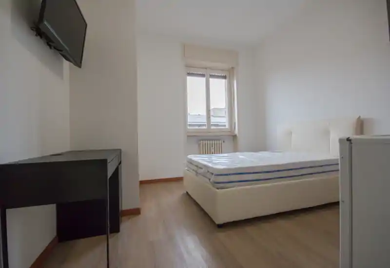 Dormitorio moderno y minimalista con cama doble y luz natural procedente de una ventana amplia; mobiliario sencillo con un pequeño escritorio y televisor en pared. El colchón sigue envuelto en plástico; suelo efecto madera y paredes blancas.