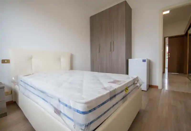 Dormitorio luminoso y minimalista con colchón limpio sobre cama tapizada crema, un armario alto de madera y suelo claro. Decoración neutra y presentación ordenada, adecuado para anuncios.