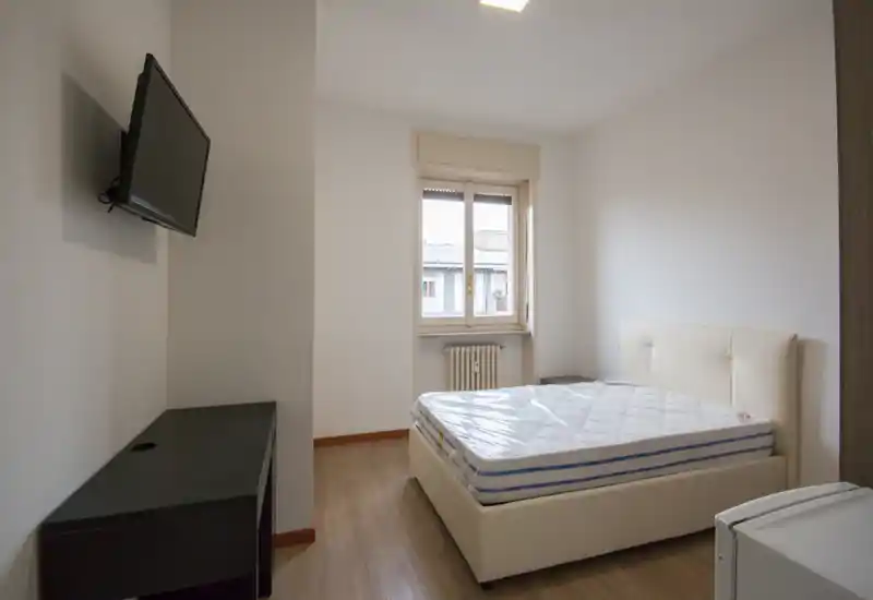 Dormitorio luminoso y minimalista con cama doble, cabecero y colchón aún en plástico. La habitación cuenta con televisor en pared, suelo de madera y una ventana que aporta luz natural.