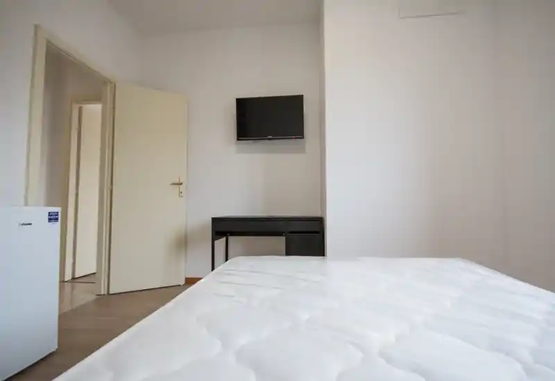 Dormitorio sencillo y minimalista con el colchón en primer plano, un pequeño escritorio y TV en la pared. Paredes blancas y suelo neutro que proporcionan una sensación luminosa y despejada, ideal para alquileres cortos.
