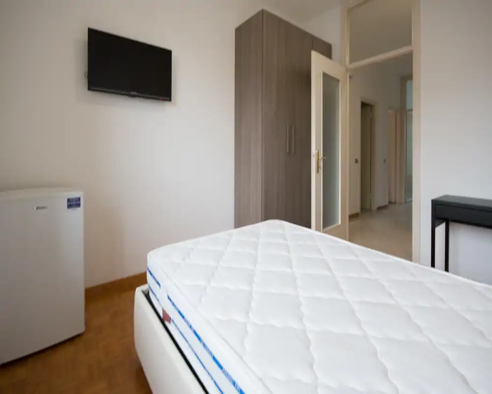 Minimal modern bedroom with a clearly visible mattress in the foreground, a wardrobe and wall-mounted TV. Bright, simply furnished space suitable for short-term rentals.