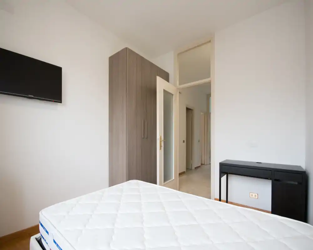 Bright, minimal bedroom with a visible mattress, wall-mounted TV and a freestanding wardrobe. Neutral tones and simple furnishings create a clean, modern feel ideal for rental listings.