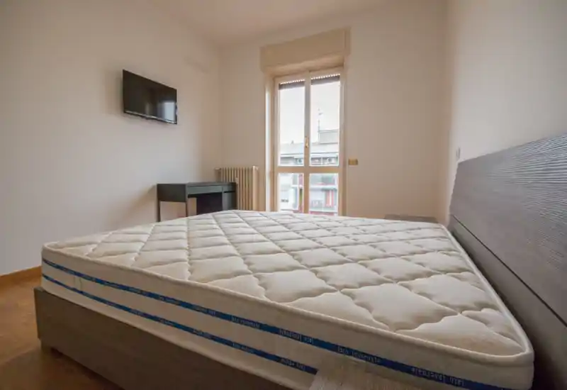 Dormitorio luminoso y sencillamente amueblado con un colchón grande y cabecero, televisor en la pared y ventana/balcón que aporta luz natural—limpio, moderno y funcional.