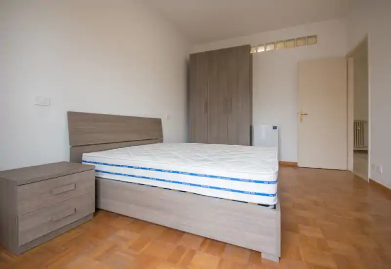 Dormitorio moderno y luminoso con cama doble amplia, mesita a juego y armario; líneas sencillas y suelo de parquet para una apariencia ordenada y lista para entrar a vivir.