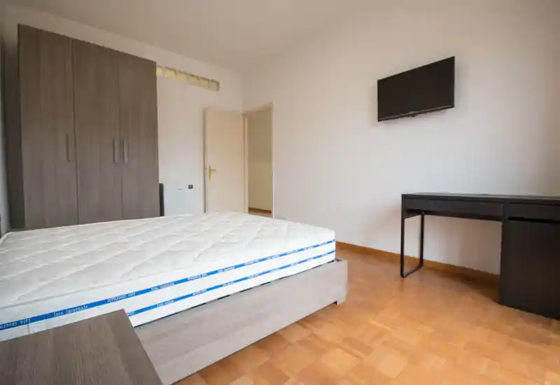 Dormitorio luminoso y minimalista con una cama grande sobre estructura de madera, armario y un escritorio con televisor en la pared: espacio limpio y funcional ideal para alquileres.