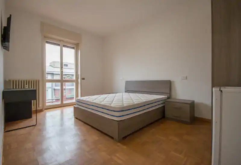 Dormitorio luminoso y sencillo con cama doble sobre estructura de madera, mesita de noche y puerta balcón que aporta luz natural.