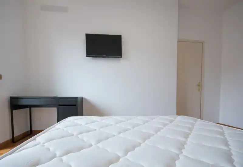 Minimal, bright bedroom with a large mattress filling the foreground, a wall-mounted TV, and a simple desk—clean, freshly prepared space ideal for a rental listing.