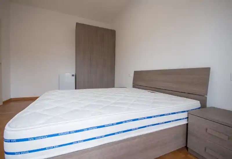 Bright, minimal bedroom with a large mattress on a wooden frame, matching headboard and bedside chest, and a wardrobe against the far wall.