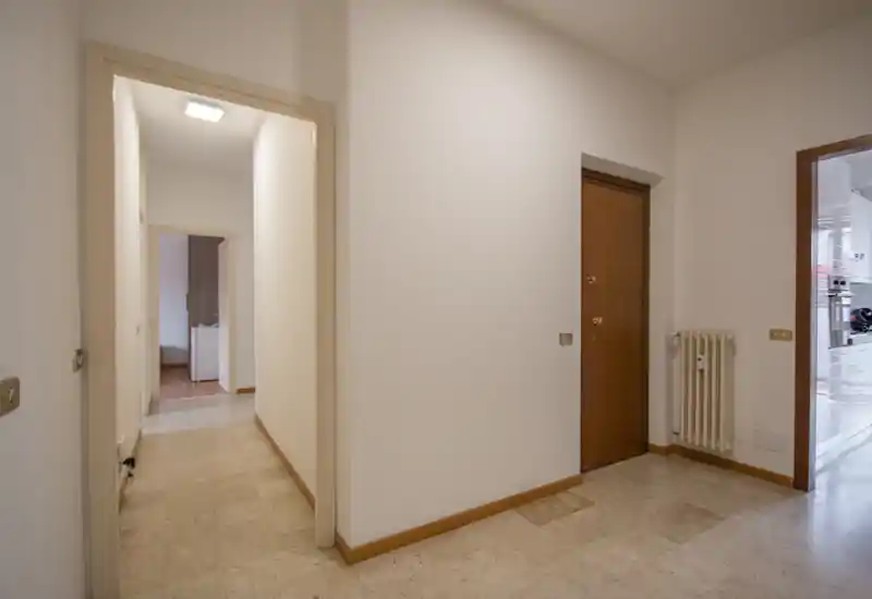 Ingresso luminoso e semplice con porta in legno, termosifone e pavimento piastrellato; pareti bianche neutre e buona visibilità verso le stanze adiacenti.