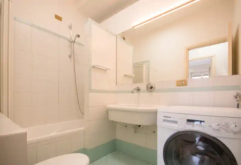 Bagno compatto e luminoso con piastrelle, vasca con doccia, lavabo a sospensione e una lavatrice a carica frontale in primo piano.