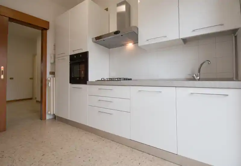 Cucina luminosa e moderna con mobili bianchi, forno integrato, piano cottura a gas e cappa in acciaio; piano di lavoro pulito con lavello incorporato.