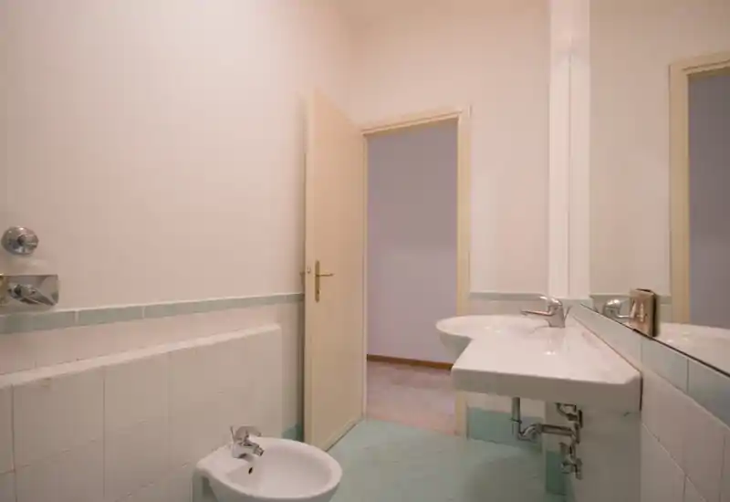 Bagno semplice e pulito con lavabo a muro, bidet e grande specchio; piastrelle pastello e illuminazione delicata creano un'atmosfera tranquilla.