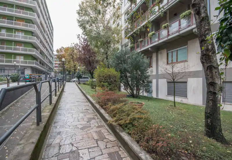 Vialetto lastricato e cortile piantumato accanto a condomini, con ringhiere e arbusti che definiscono un'area esterna comune ordinata.