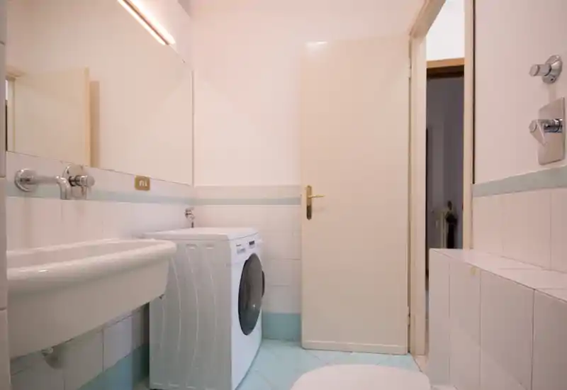 Bagno compatto e pulito con lavabo, specchio a parete e una lavatrice a carica frontale. Piastrelle chiare e illuminazione soffusa creano uno spazio ordinato e funzionale.