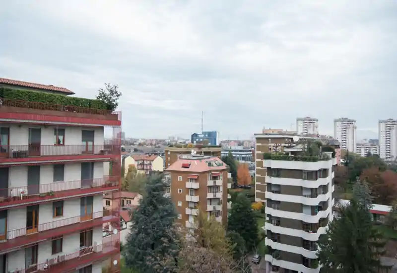 Ampia vista esterna da un piano alto con palazzi vicini, alberi e lo skyline cittadino in lontananza — ideale per descrivere la vista da un balcone o terrazza.