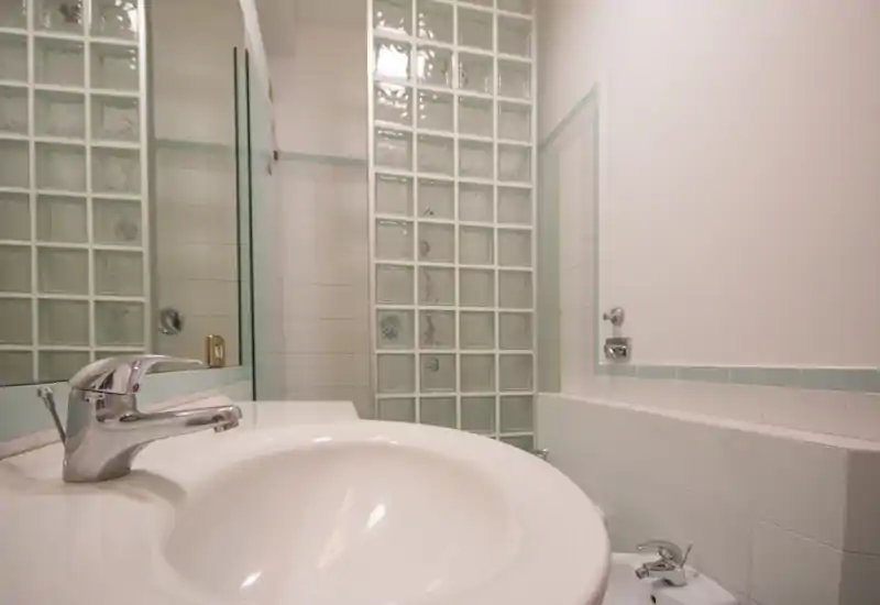 Area lavabo di un bagno moderno e luminoso con rubinetto cromato, piastrelle e una parete in vetrocemento che garantisce privacy e ingresso di luce.