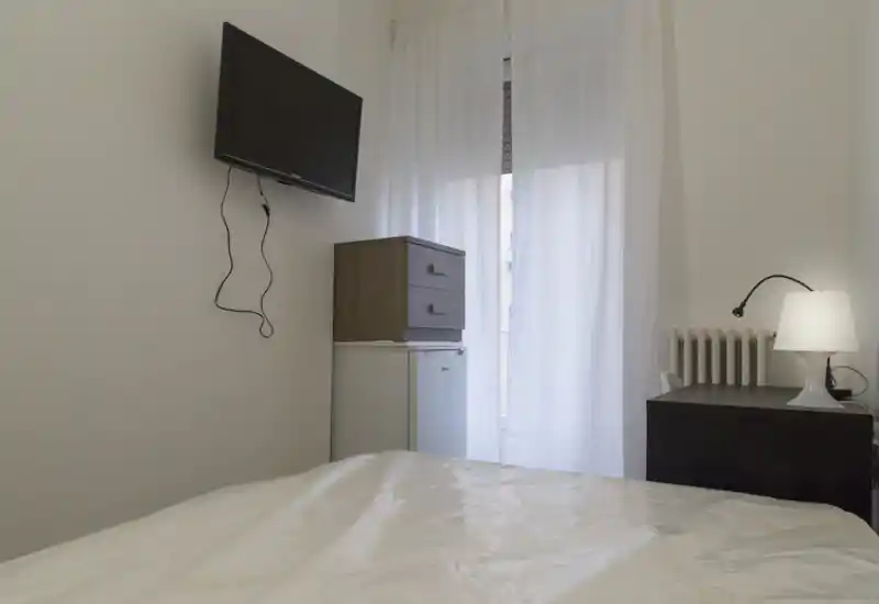Simple bedroom view with a visible bed foreground, wall-mounted TV, small dresser over a mini-fridge and a bedside table with lamp; sheer curtains admit soft natural light.