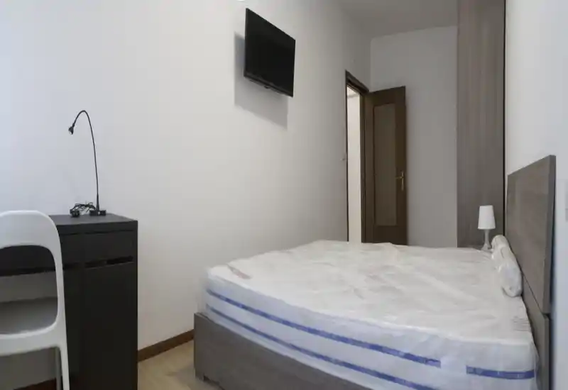 Chambre moderne et sobre avec un lit double (matelas encore sous plastique), une tête de lit, une lampe de chevet, un bureau avec lampe et une TV murale — décor lumineux et neutre, idéal pour les locations courtes durées.